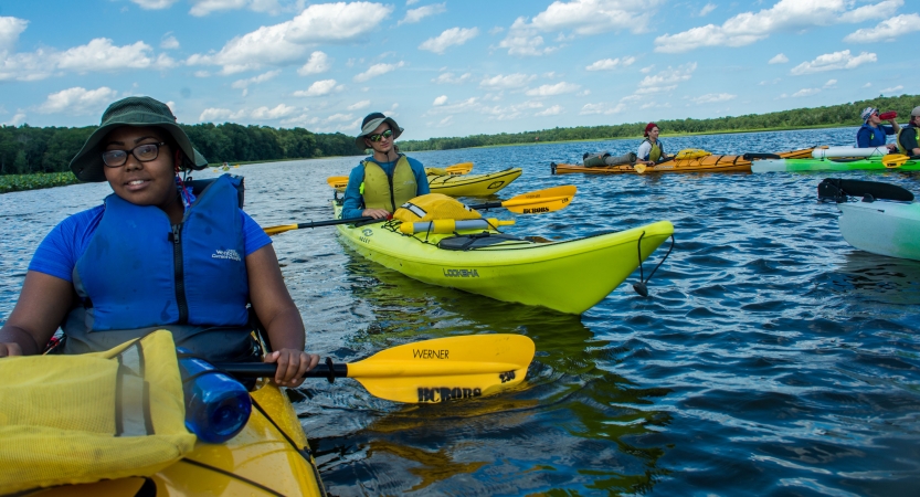 a group of teens take a break from paddling kayaks on a large body of water lined by trees